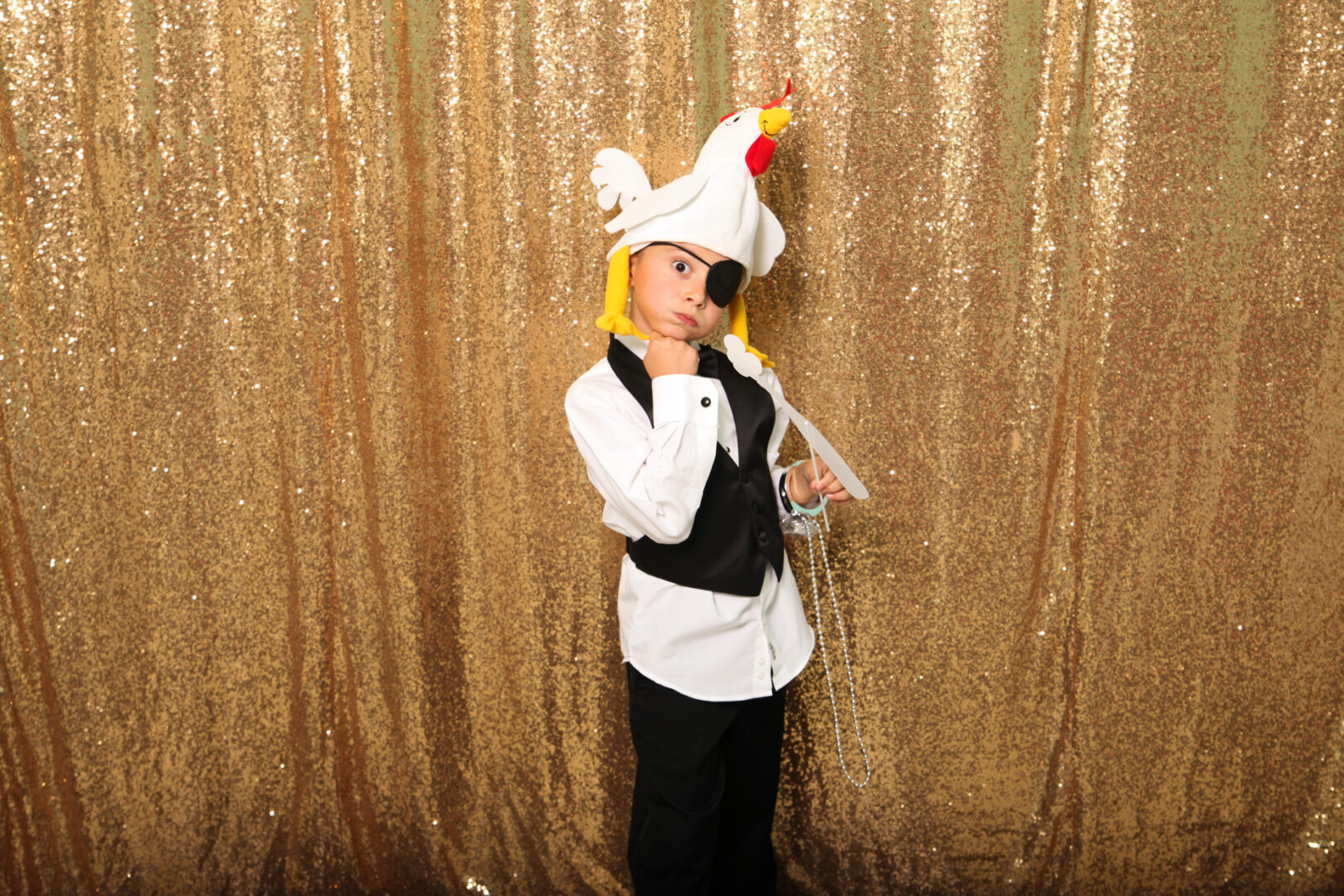 A child in a black vest and white shirt poses against a gold sequined backdrop in the Cape May Photo Booth, wearing a hat with a stuffed chicken on top.
