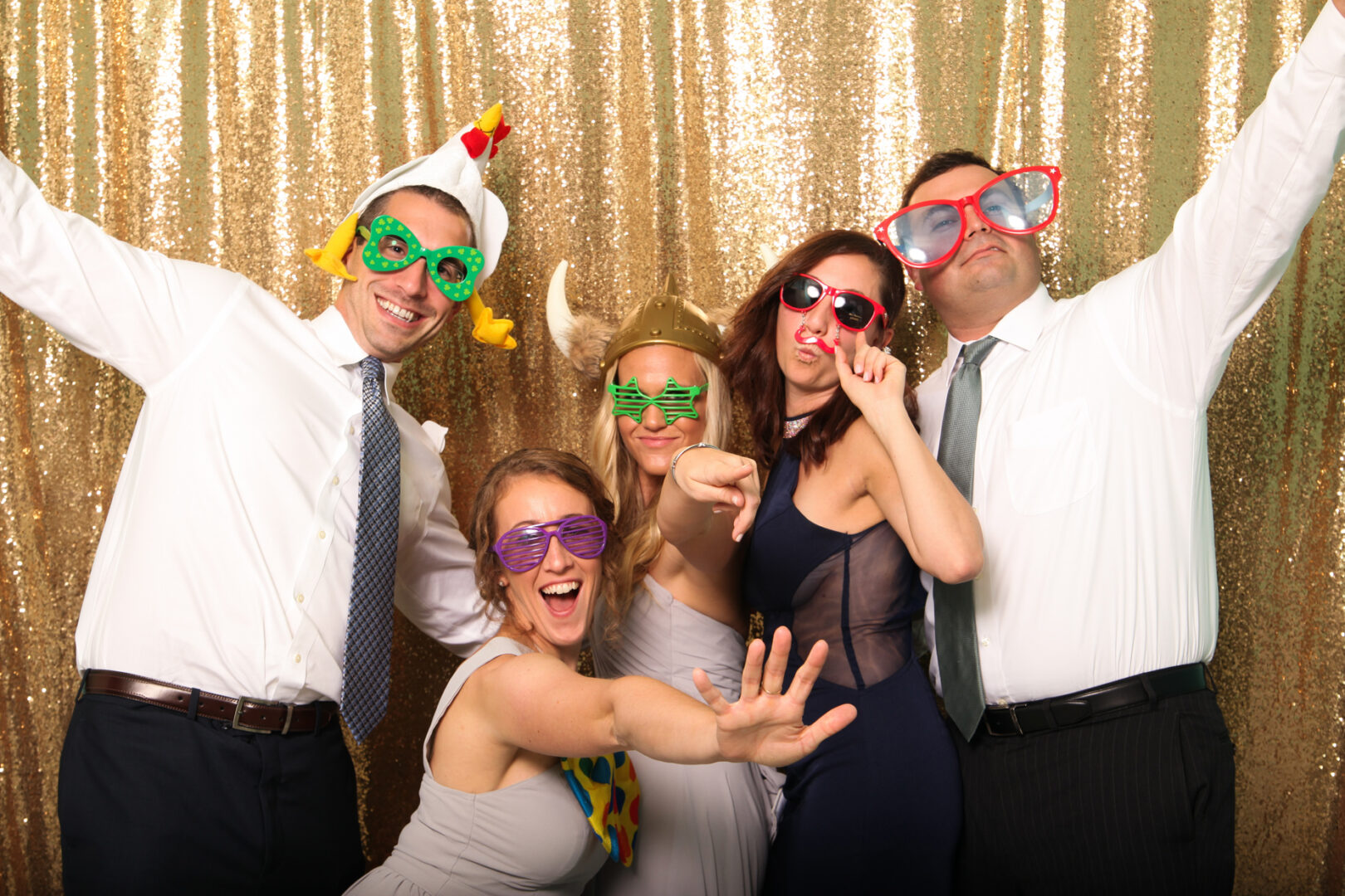 Five people wearing playful costumes and colorful glasses pose energetically in front of a gold sequin backdrop at the Cape May Photo Booth.