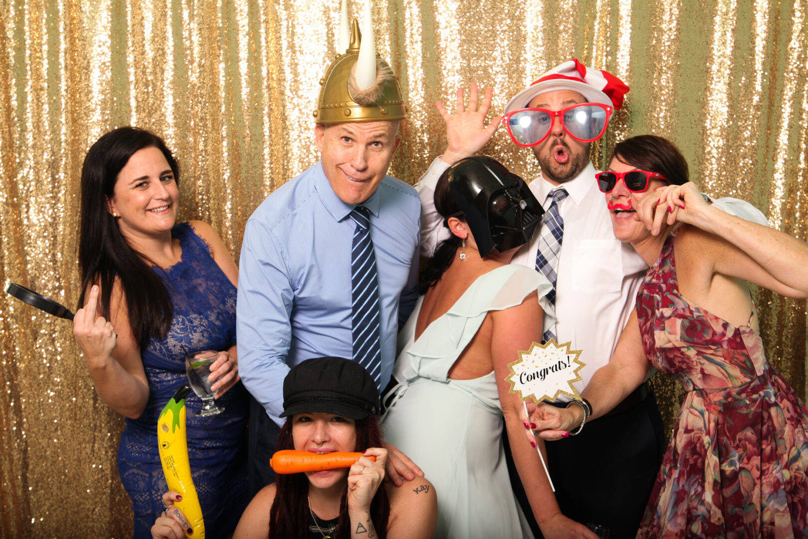 A group of six people poses in front of a gold sequin backdrop at the Cape May Photo Booth, donning various hats and novelty glasses while playfully holding props like a banana and carrot.