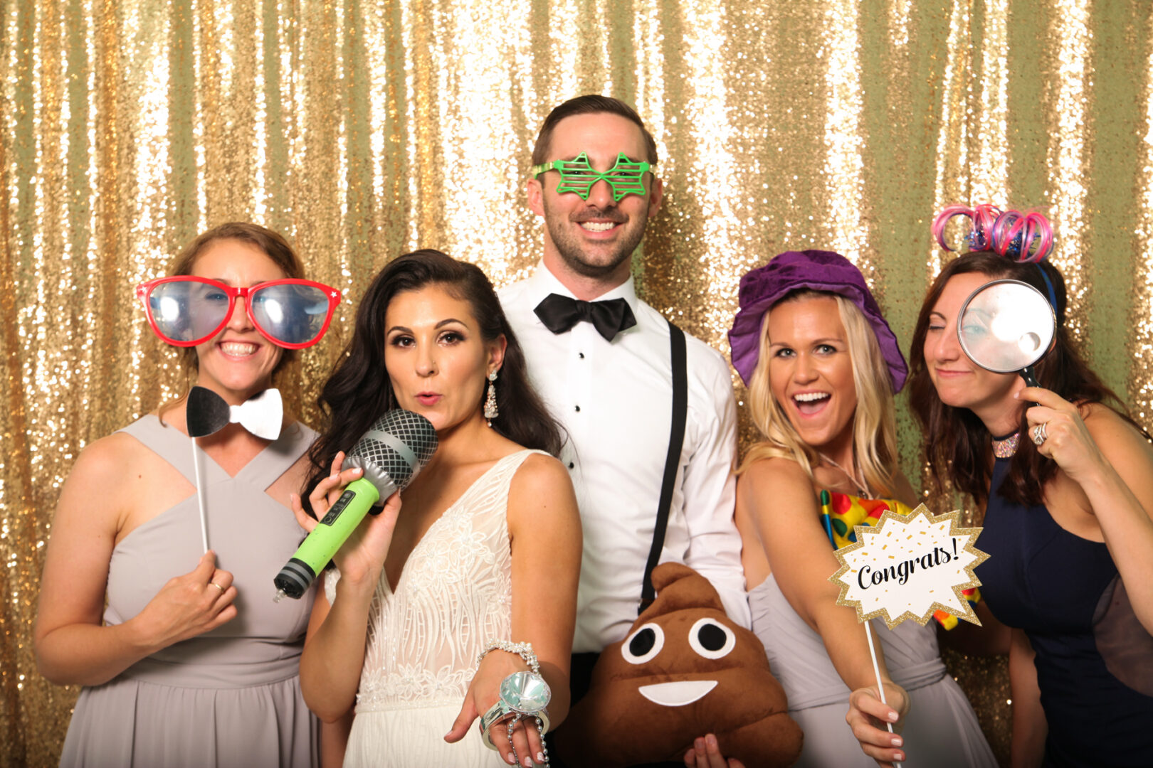 Five people pose in front of a gold sequin backdrop, decked out with playful props like oversized glasses, a microphone, and a "Congrats" sign. It's the perfect memory captured by Cape May Photo Booth.