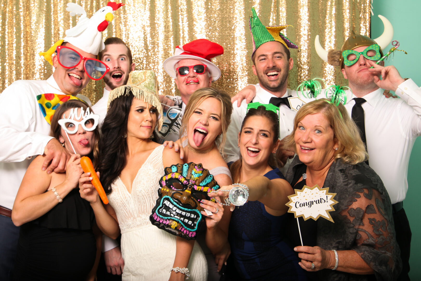 A lively group poses energetically before a gold sequin backdrop at Cape May Photo Booth, donning playful hats like a Viking helmet, silly glasses, and holding whimsical signs and toys.