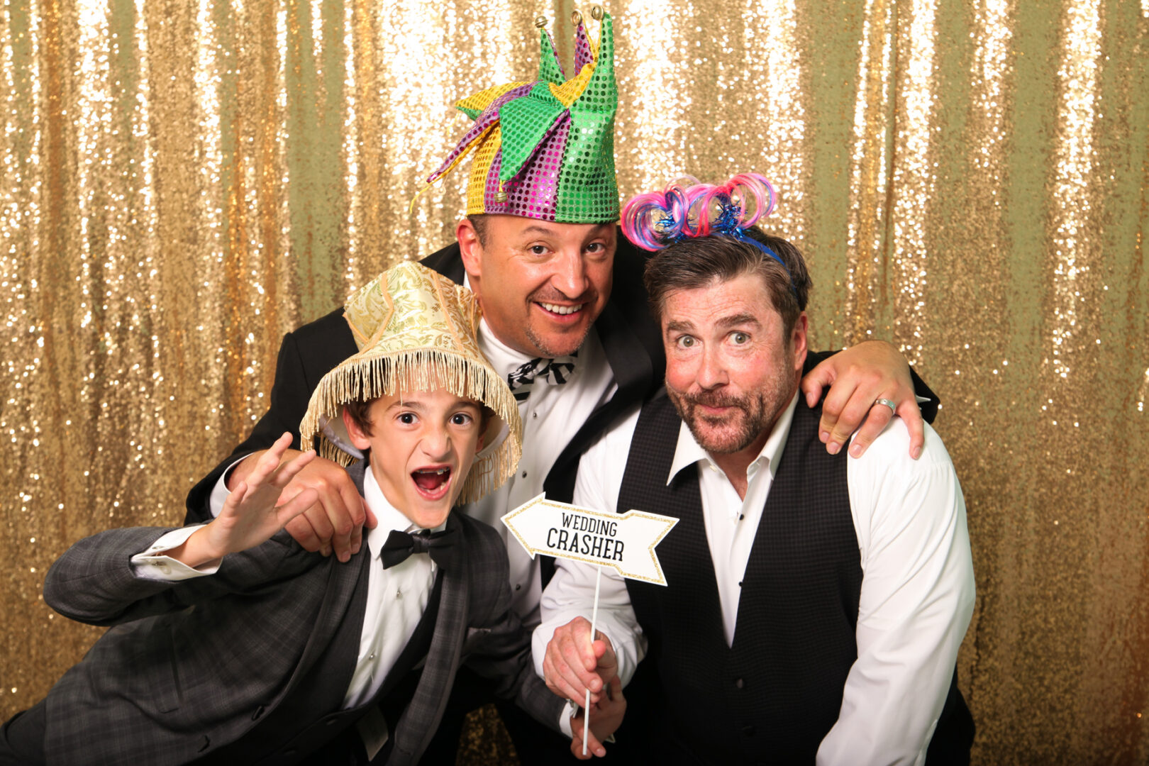 Three men in suits and festive hats pose in front of a gold sequin backdrop from Cape May Photo Booth. One holds a "Wedding Crasher" sign, smiling.