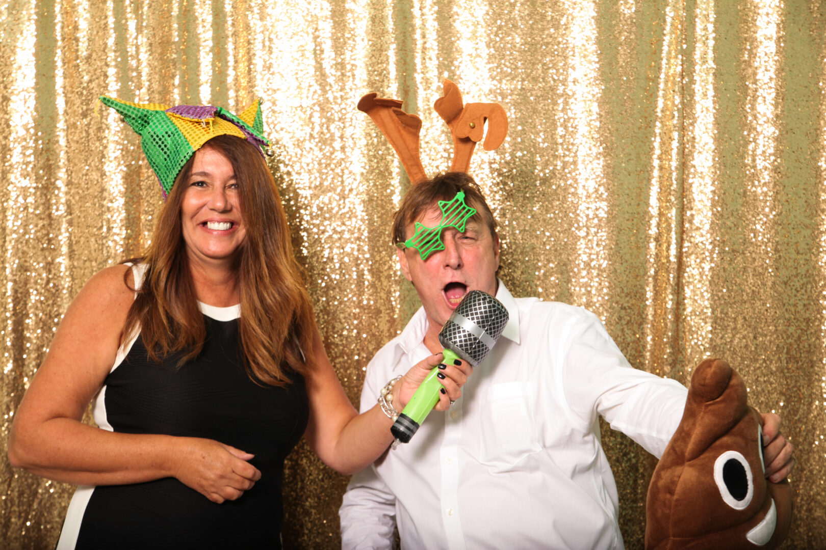 Two people pose joyfully in front of a gold sequin backdrop provided by Cape May Photo Booth. They're sporting festive hats and novelty glasses; one wields a toy microphone, the other clutches a plush emoji, capturing the spirit of fun and celebration.
