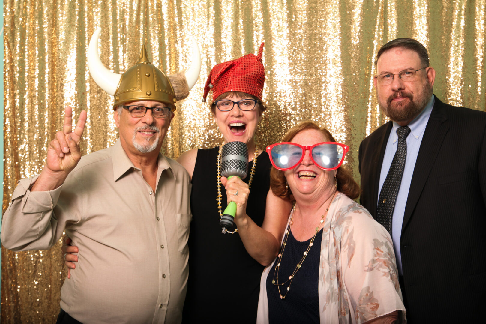 Four people posing in front of a gold sequin backdrop at the Cape May Photo Booth. One wears a Viking helmet, another a red hat and holds a toy microphone, the third sports oversized glasses, and the fourth is looking sharp in a suit.