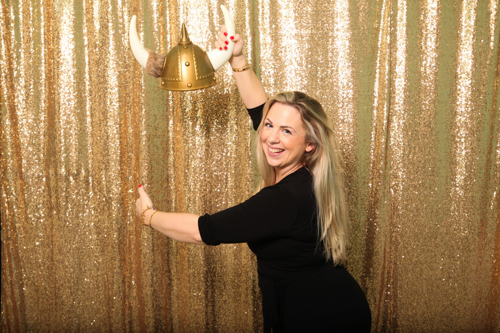 A person in a black outfit smiles and poses with a Viking helmet in front of a glittery gold curtain at the Cape May Photo Booth.