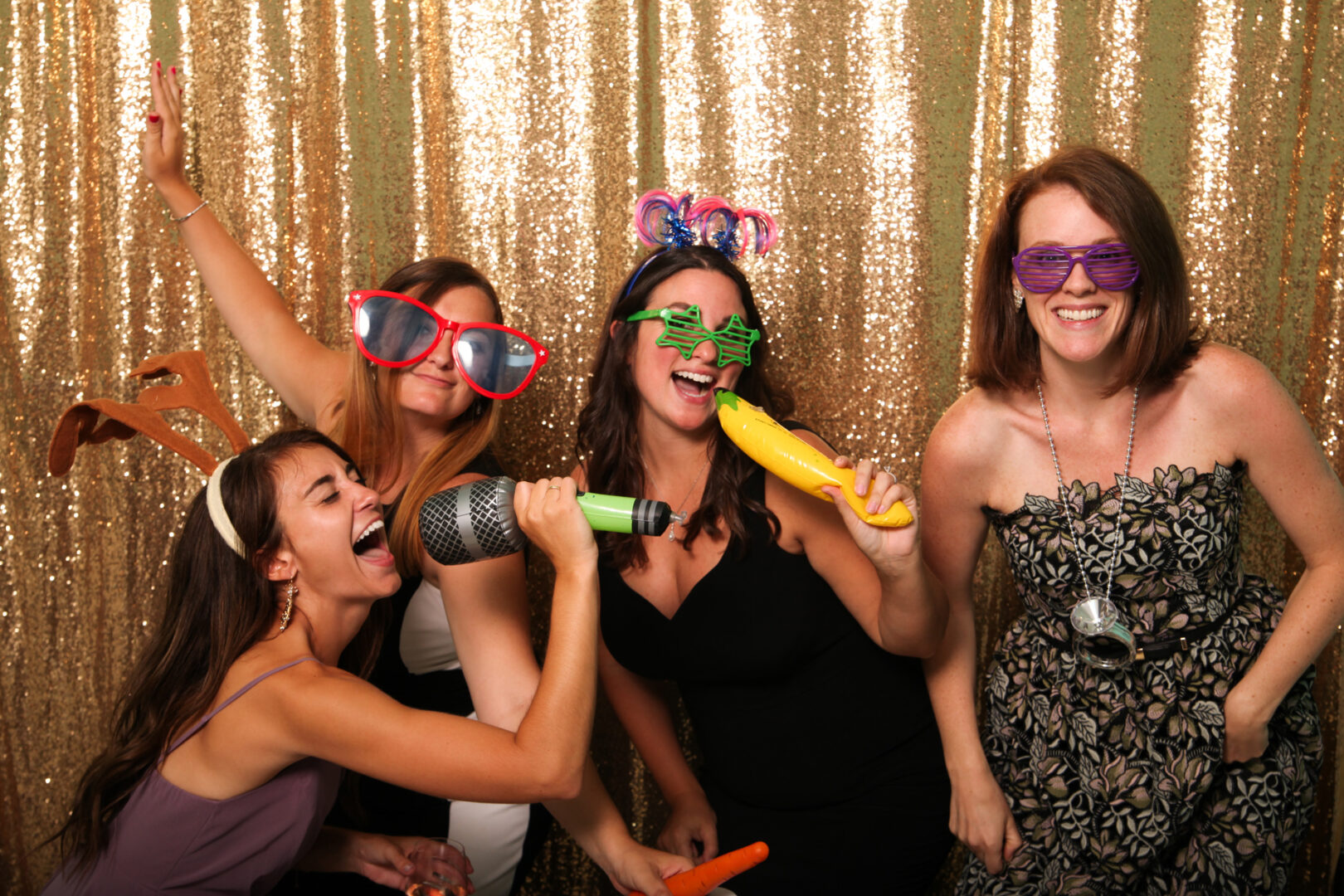 Four women in a Cape May Photo Booth strike playful poses with oversized glasses, novelty microphones, and quirky headbands against a shimmering gold sequin backdrop, capturing the joy of the moment.
