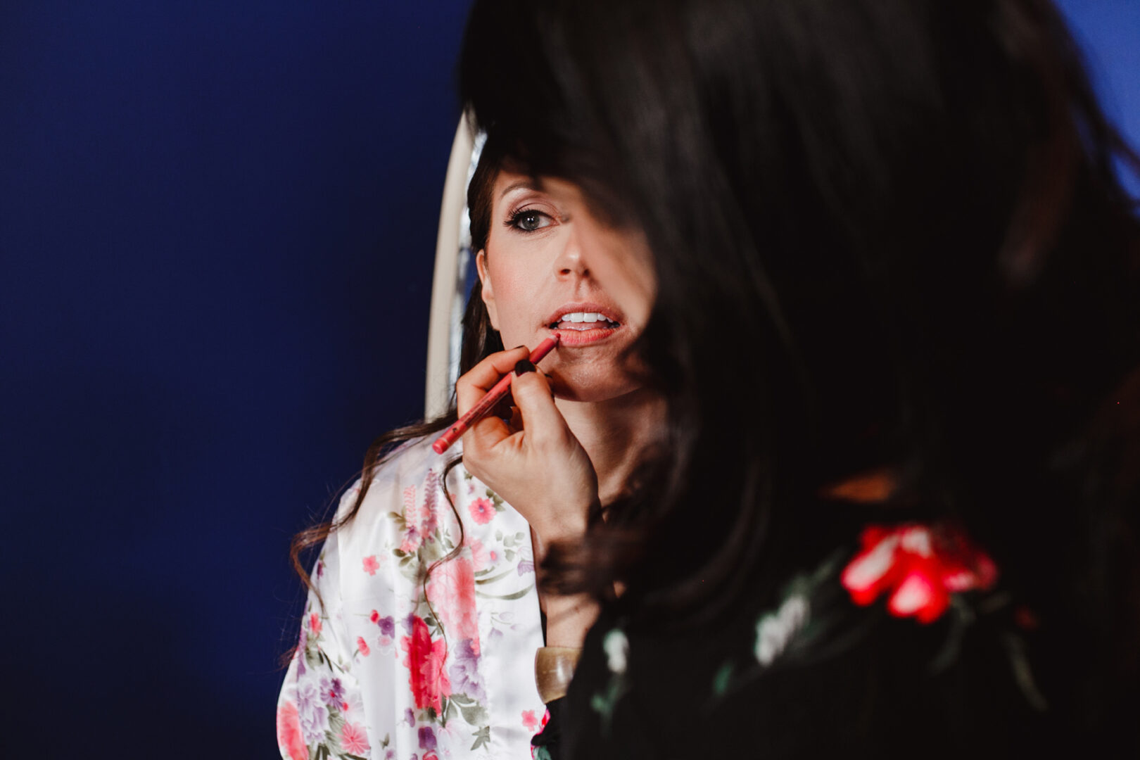 A woman with dark hair applies lip makeup to another woman wearing a floral robe, using a lip liner, in preparation for a Cape May wedding. The background is a deep blue.