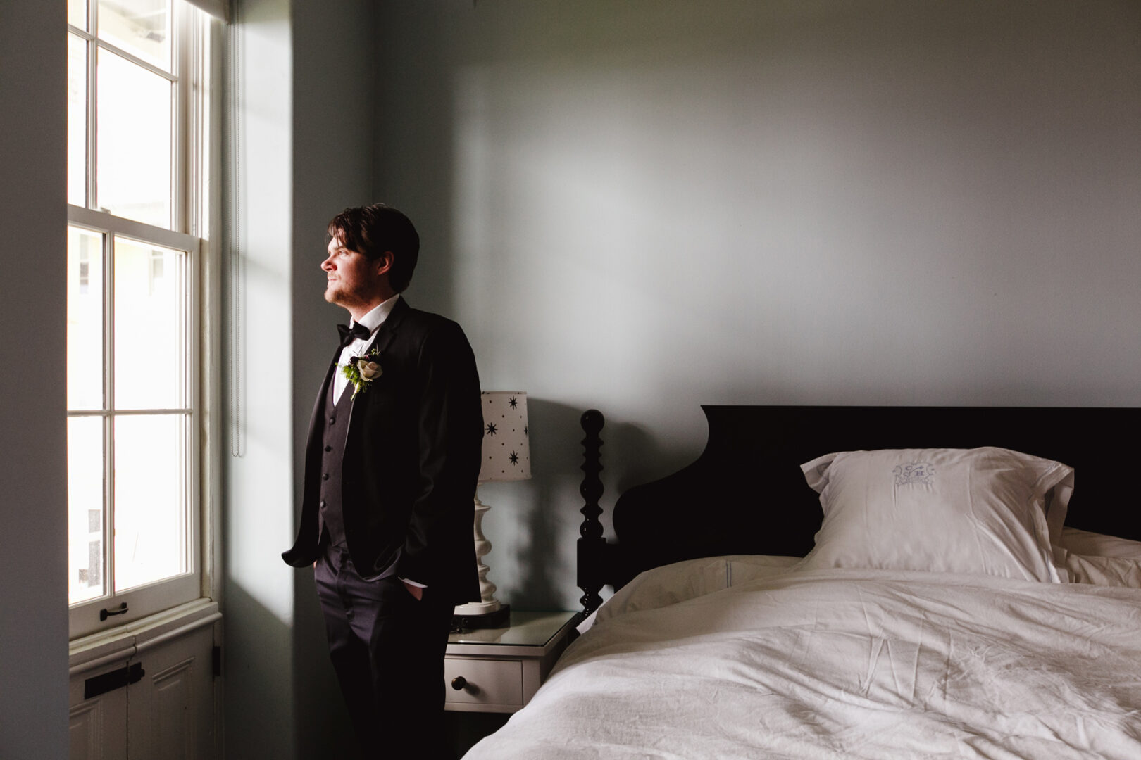A man in a tuxedo stands by a window, gazing thoughtfully at the serene Cape May wedding outside. A bed and nightstand are visible in the room, adding a cozy touch to this special day.