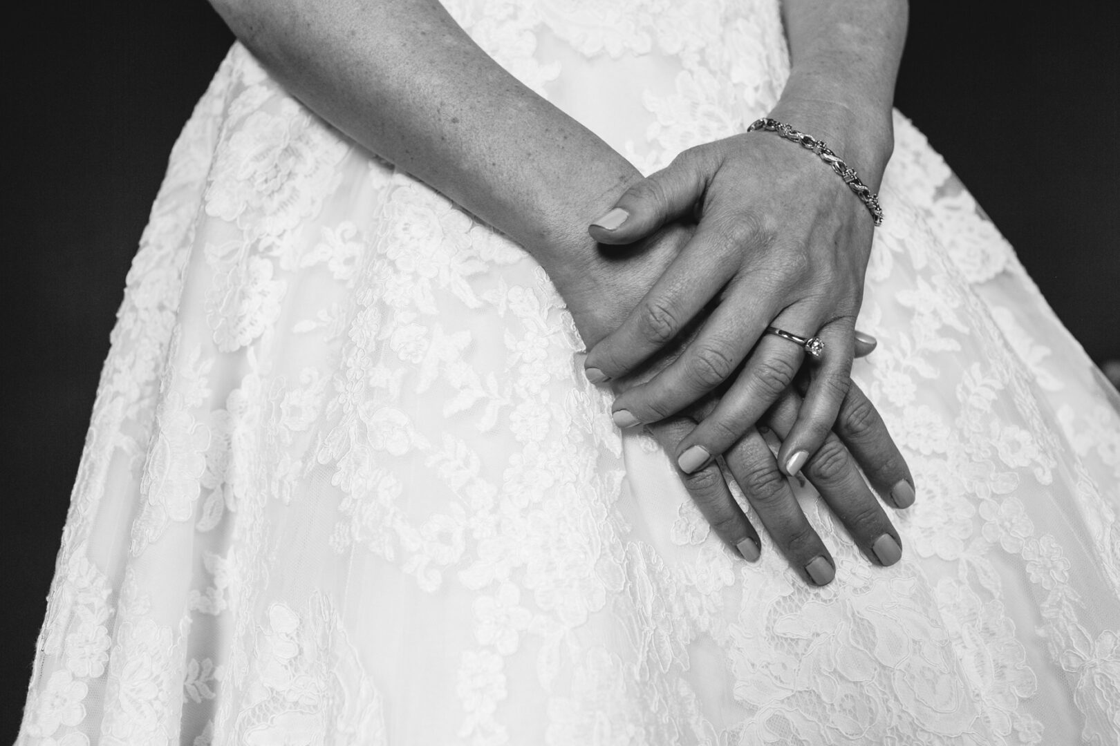 A close-up of a person at a Cape May wedding, clad in a white lace dress with hands elegantly clasped, showcasing both a bracelet and a ring on the finger.