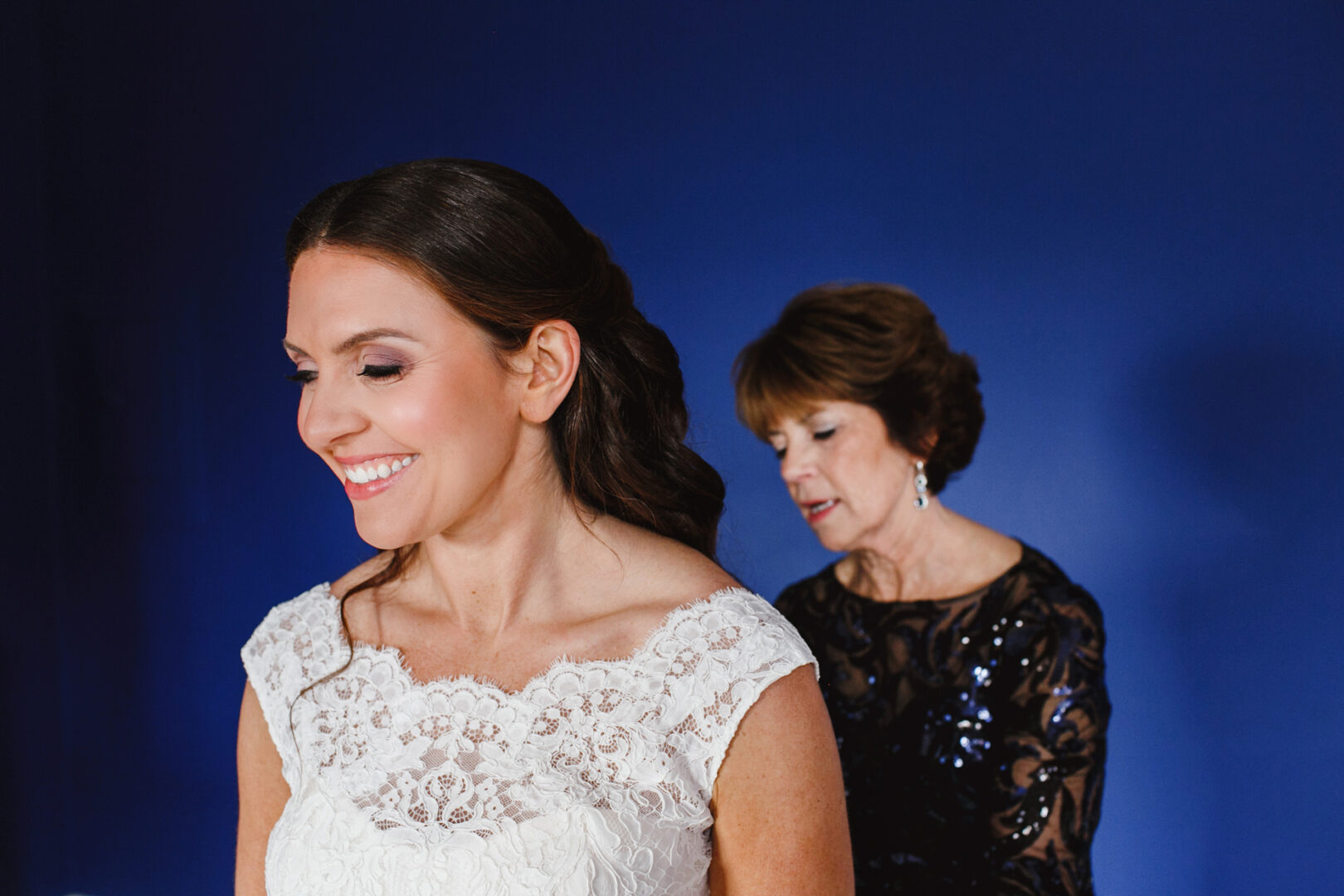 A woman in a lace bridal dress smiles radiantly, embodying the essence of a Cape May wedding, while another woman in a dark sequined dress stands gracefully behind her against a solid blue backdrop.