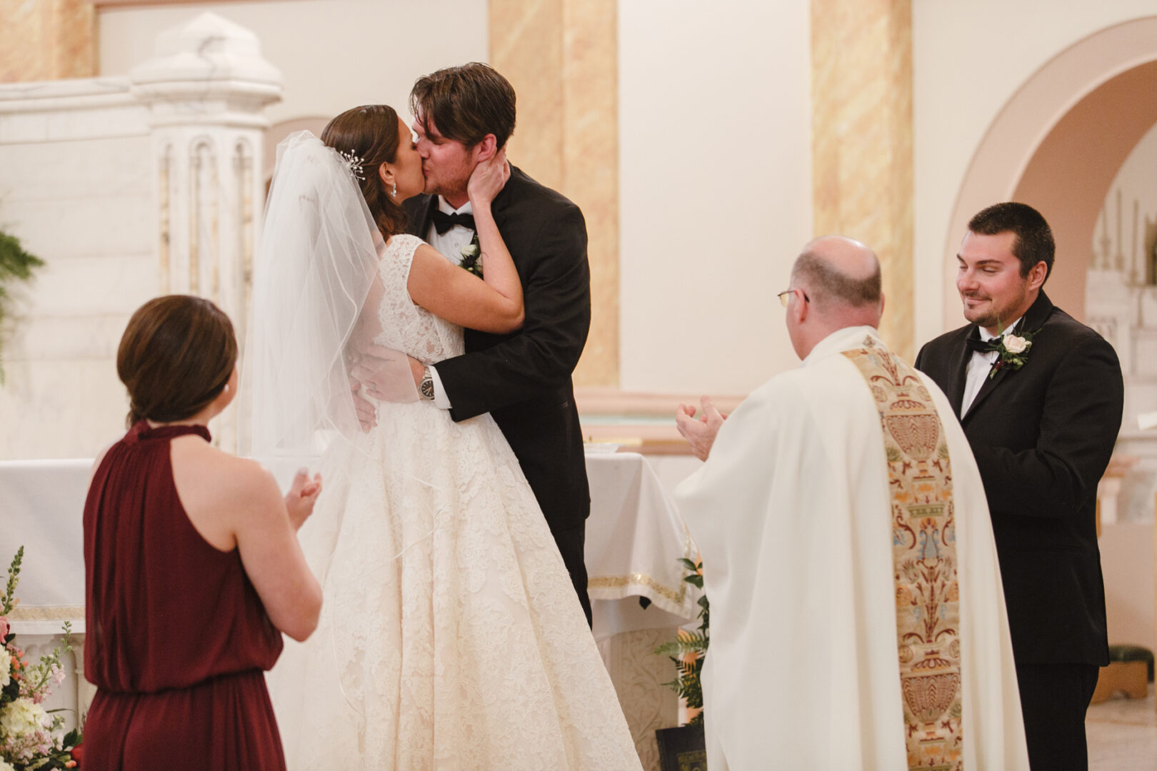 At a charming Cape May wedding, a bride and groom share a tender kiss at the altar. A priest and two attendees stand nearby, capturing the magic of the moment.