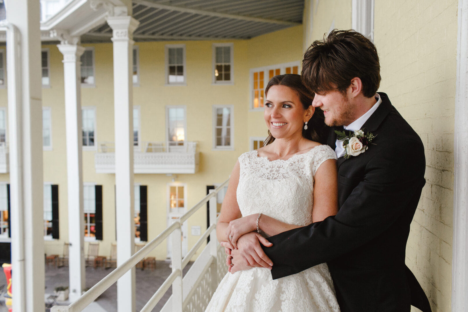 On a charming Cape May wedding balcony, a groom embraces his bride from behind. She is stunning in her white gown, and he looks dapper in his black suit. They stand gracefully before a yellow building adorned with elegant white columns.
