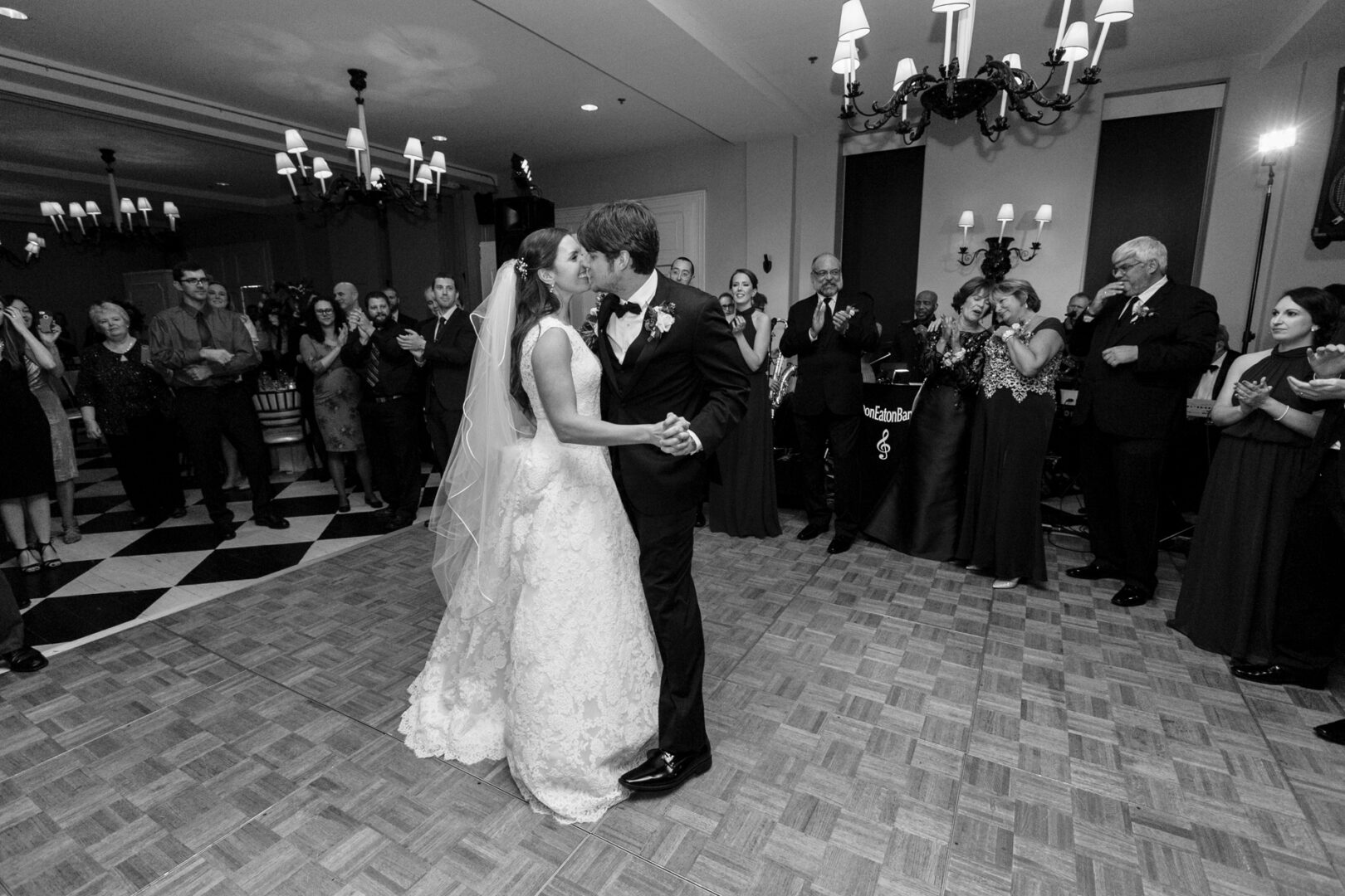 In a stunning Cape May wedding, the bride and groom share their first dance in a ballroom, surrounded by guests clapping and smiling. The elegant setting features chandeliers and a checkered floor, creating an unforgettable moment.