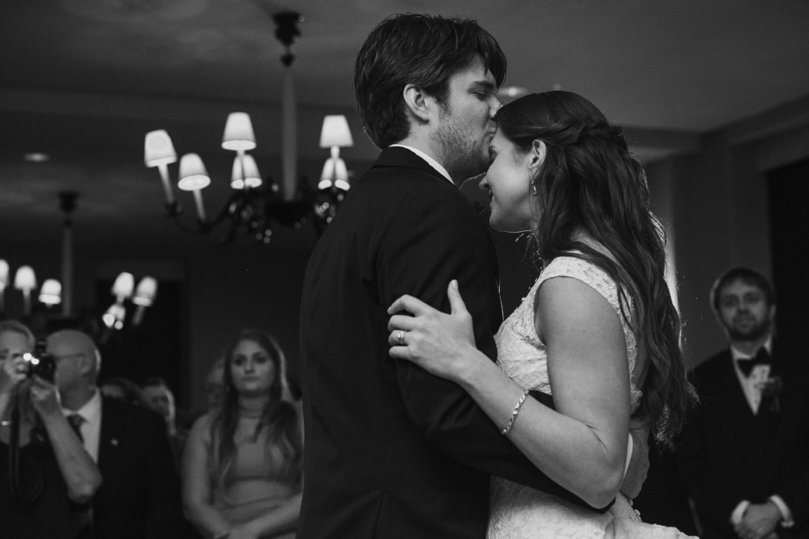 At a Cape May wedding, a couple dances closely in formal attire under chandelier lights, with guests mingling in the background.