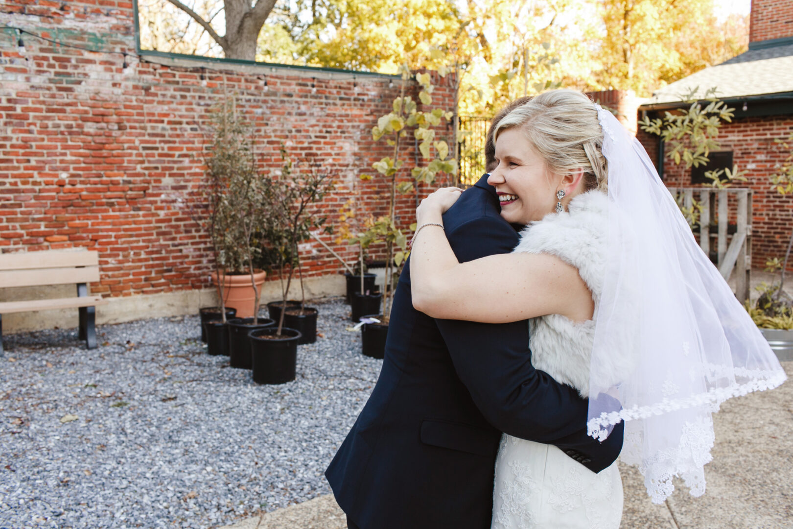 At a beautiful Delaware Center for Horticulture wedding, the bride in her white dress and veil embraces a loved one in a dark suit. The charming backdrop features a rustic brick wall and lush potted plants, setting the perfect scene for this heartfelt moment.