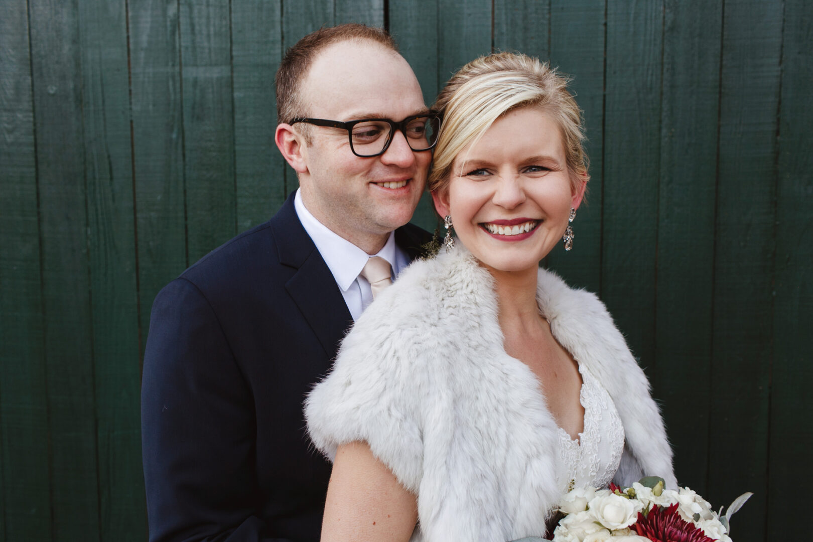 A couple poses in elegant formal attire; the woman, holding a bouquet, wears a fur stole, while the man in glasses dons a sharp suit. They stand gracefully before the dark green wooden wall, capturing a serene moment at their Delaware Center for Horticulture wedding.