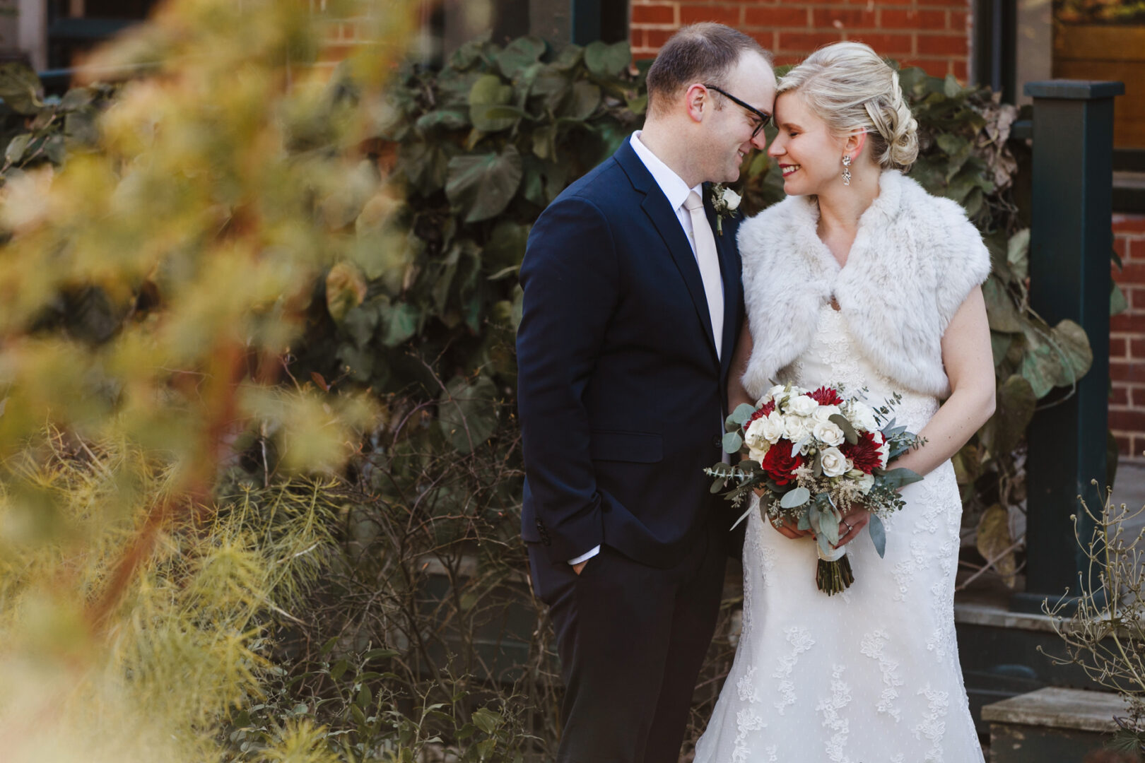 A couple stands close, smiling at each other amid the lush backdrop of the Delaware Center for Horticulture. The woman holds a bouquet of red and white flowers, wearing a white dress with a fur wrap. The man, in a dark suit, beams by her side under the serene greenery.