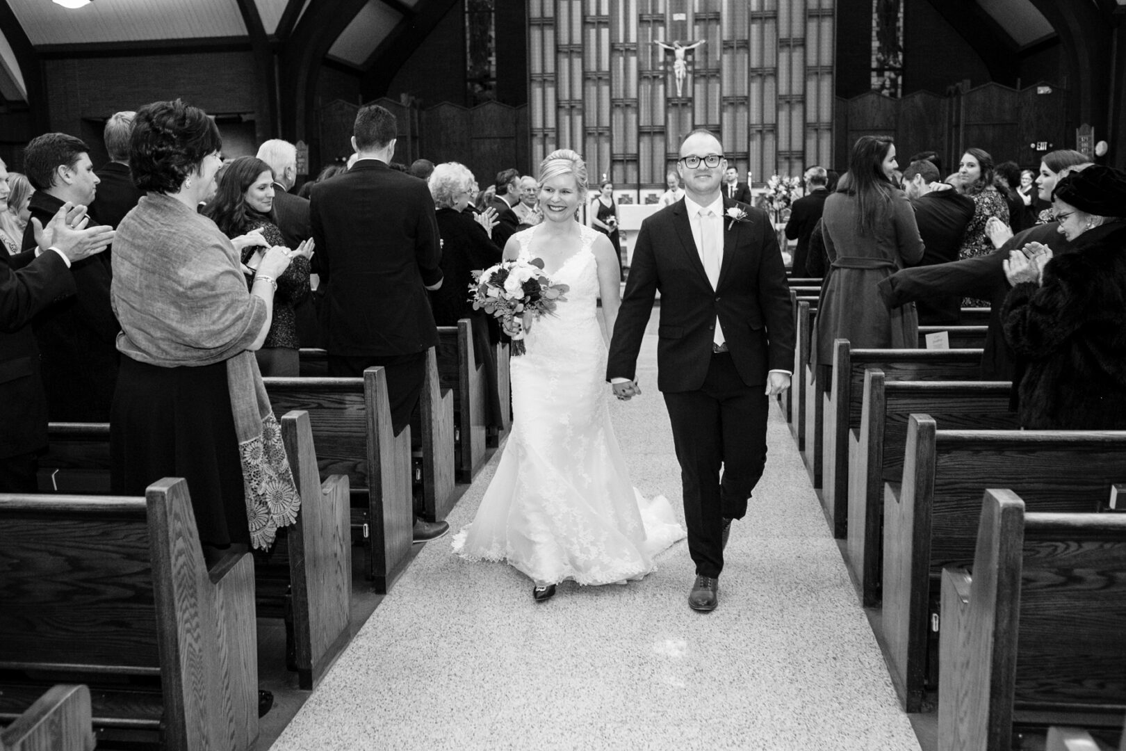 A bride and groom stroll down the aisle of a church, hand in hand, as guests clap and smile. The groom dons a suit while the bride, in a white gown with a bouquet, radiates joy at this Delaware Center for Horticulture wedding.