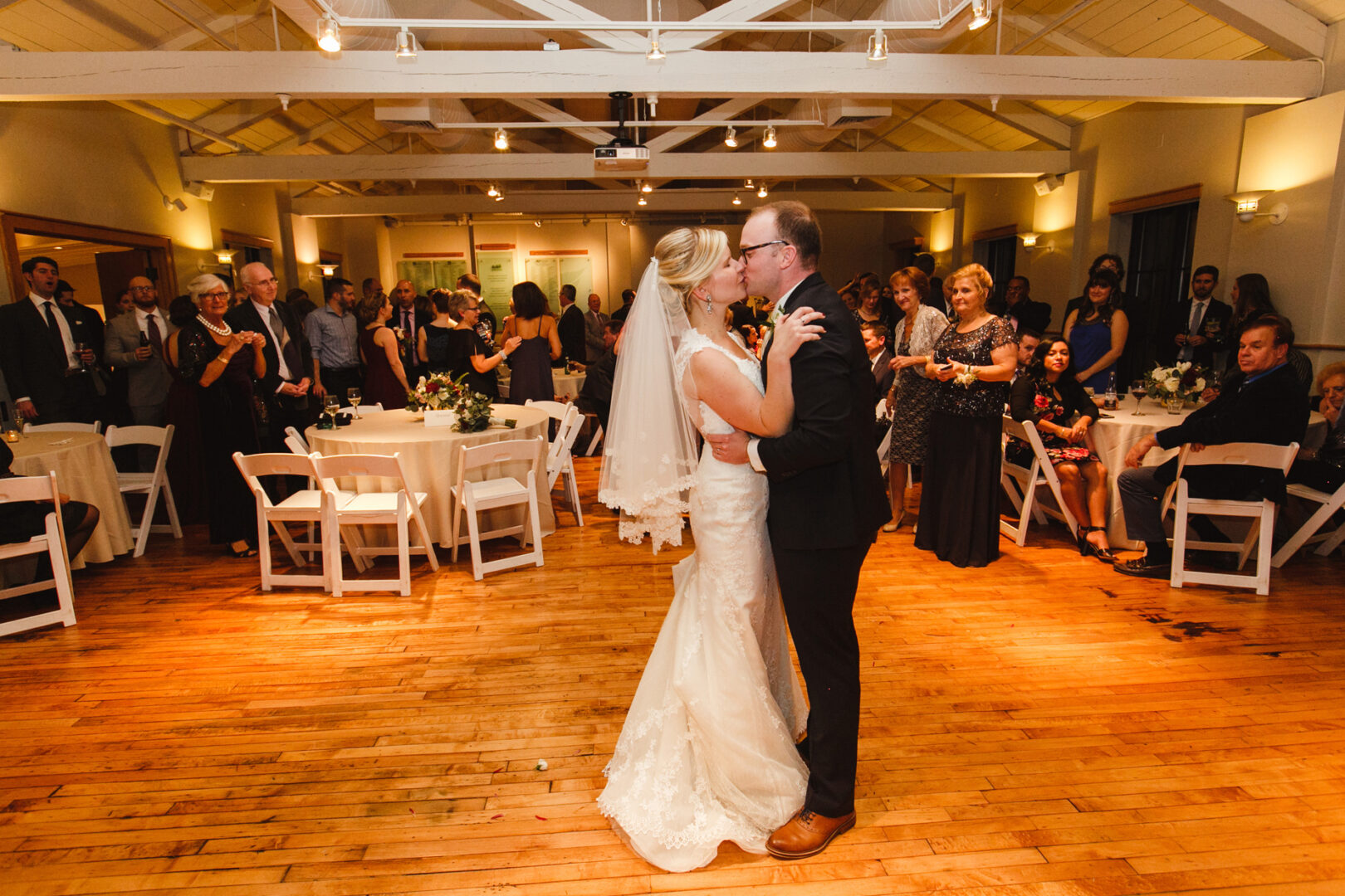 At the Delaware Center for Horticulture, a bride and groom share their first dance amid lush greenery. Wooden floors gleam under soft lights as guests seated at round tables watch the magical moment unfold in this enchanting wedding setting.