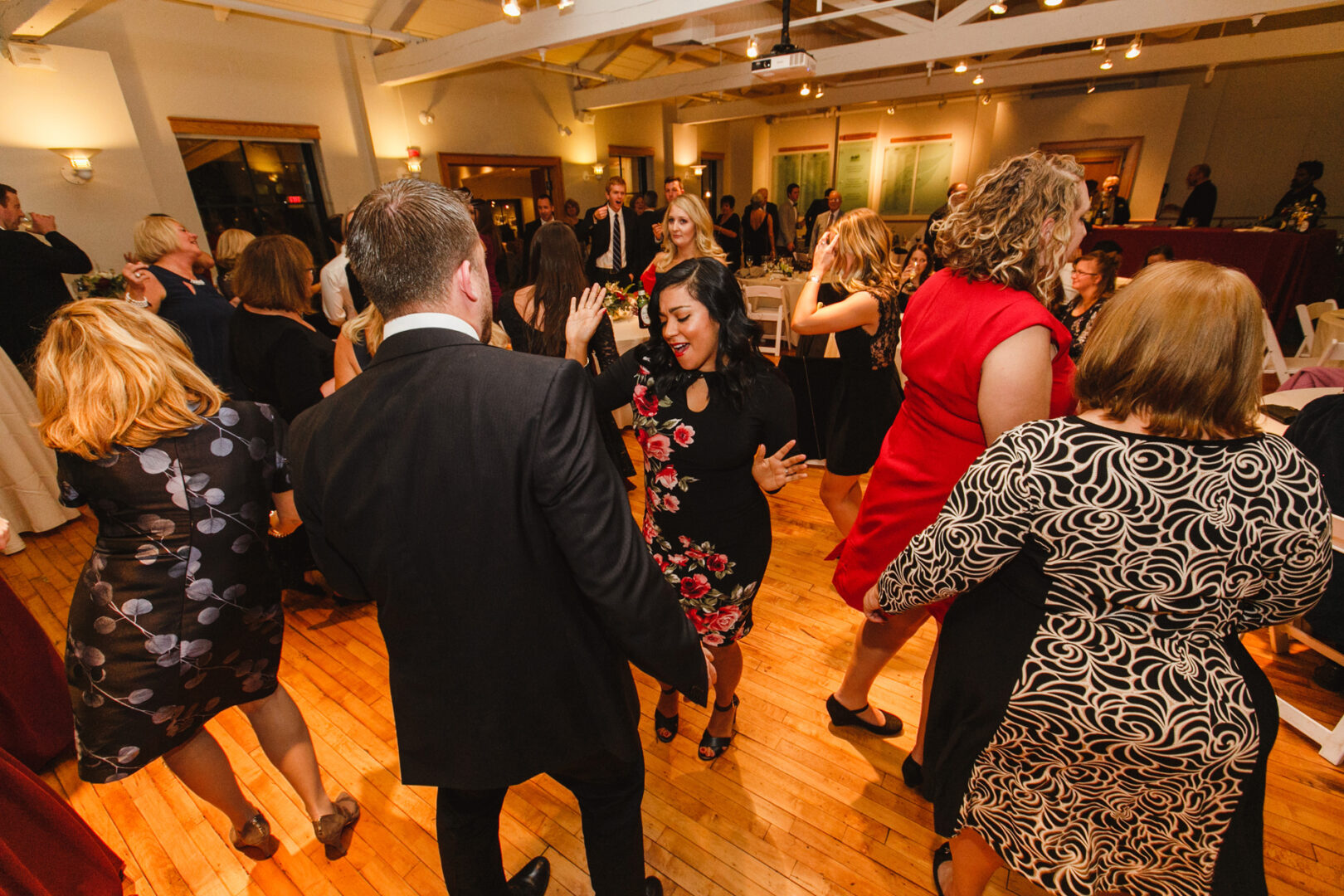 At the Delaware Center for Horticulture, guests in formal attire danced energetically at a vibrant wedding celebration, set against the charming backdrop of a well-lit indoor space with gleaming wooden floors.