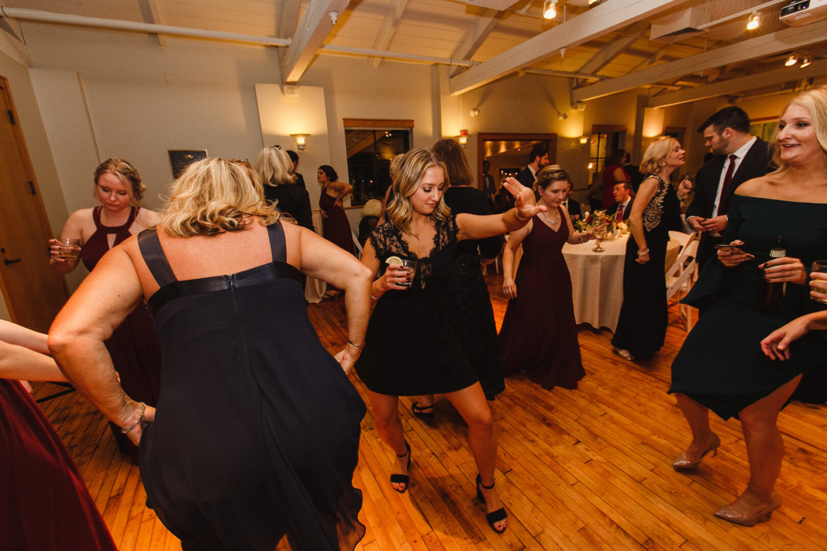 People are dancing and socializing in a warmly lit room with wooden floors at the Delaware Center for Horticulture wedding. Some hold drinks, while tables adorned with flowers grace the background, creating a charming atmosphere.