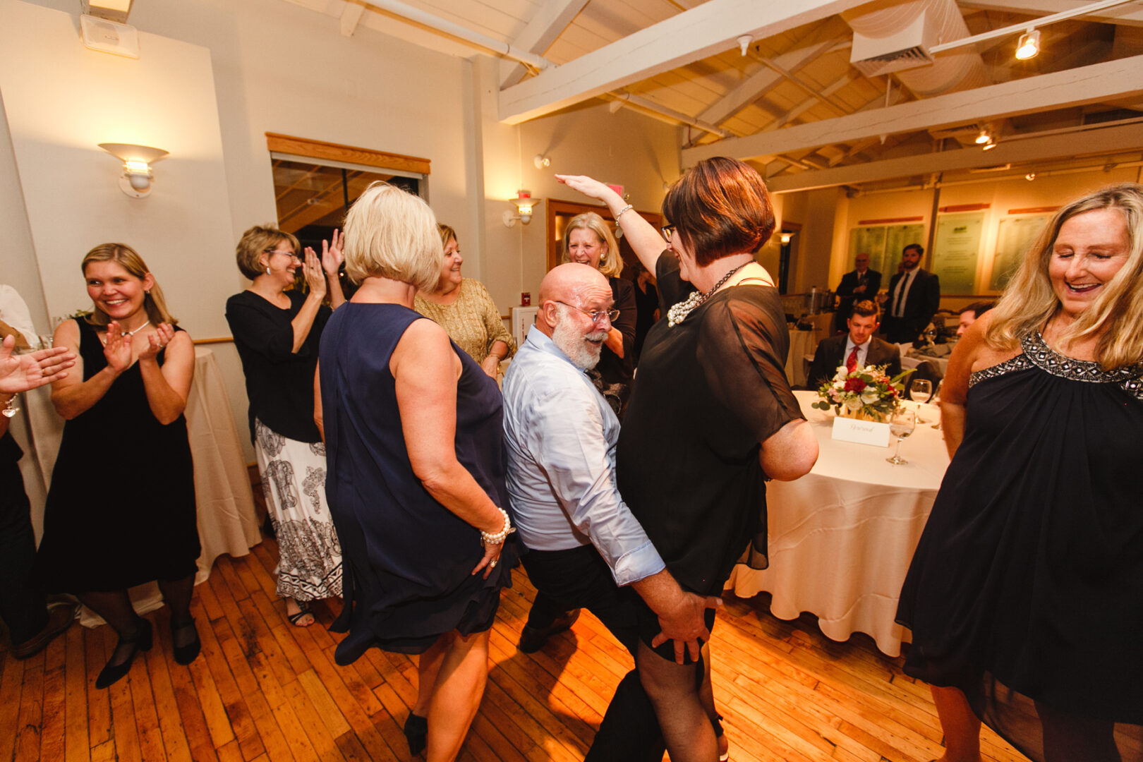 A group of people dancing in a warmly lit room with wooden floors at the Delaware Center for Horticulture. A bearded man in the center leans forward while others around him are clapping and smiling, celebrating a joyous wedding event.