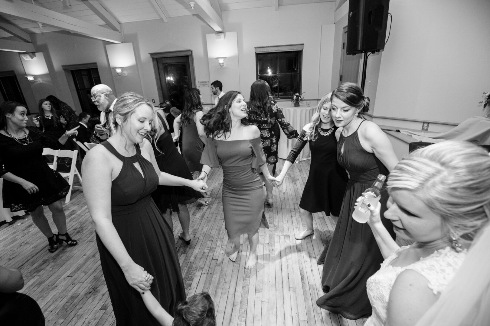 Guests in formal attire dance in a circle on the wooden floor, celebrating a beautiful wedding at the Delaware Center for Horticulture. The light-colored room adds an airy elegance to the joyful gathering.