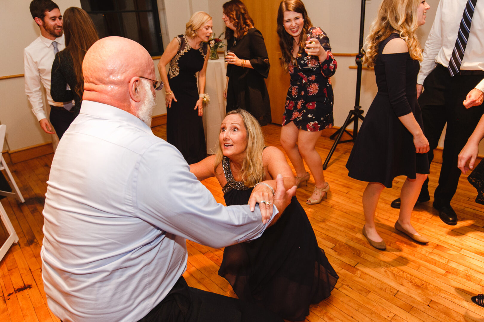 At a Delaware Center for Horticulture wedding, guests danced and socialized on the wooden floor. A bald gentleman kindly assisted a woman up from a crouch amidst the lively celebration.