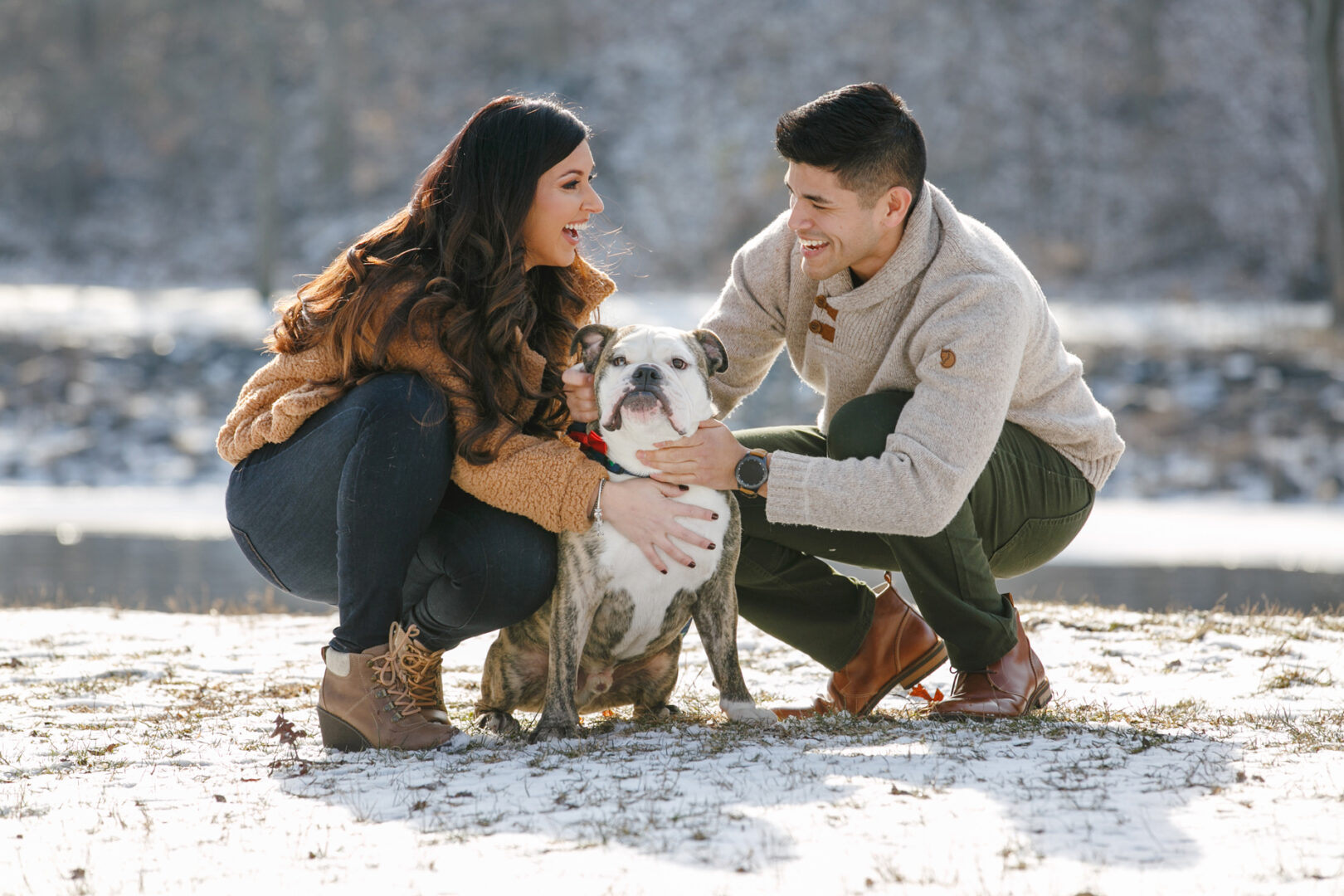 A couple beams with joy during a New Hope engagement session, crouching beside their sitting bulldog on a snowy field. Dressed warmly in winter clothes, they capture a moment of love and happiness amid the serene landscape.