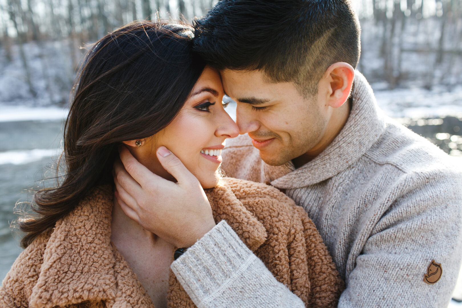 A couple in warm clothing embraces by a snowy riverbank, captured in their New Hope engagement session. The man gently holds the woman's face as they both smile and gaze at each other, with trees softly blurred in the background.