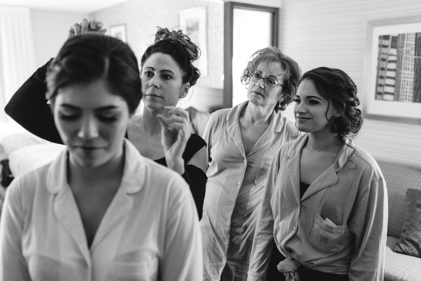 Four women in a cozy room, clad in pajamas, share a tranquil moment. Three stand behind a seated woman, one expertly styling her hair with the calm grace of a bride at the Palace at Somerset Park wedding preparations. The scene is serene and focused.