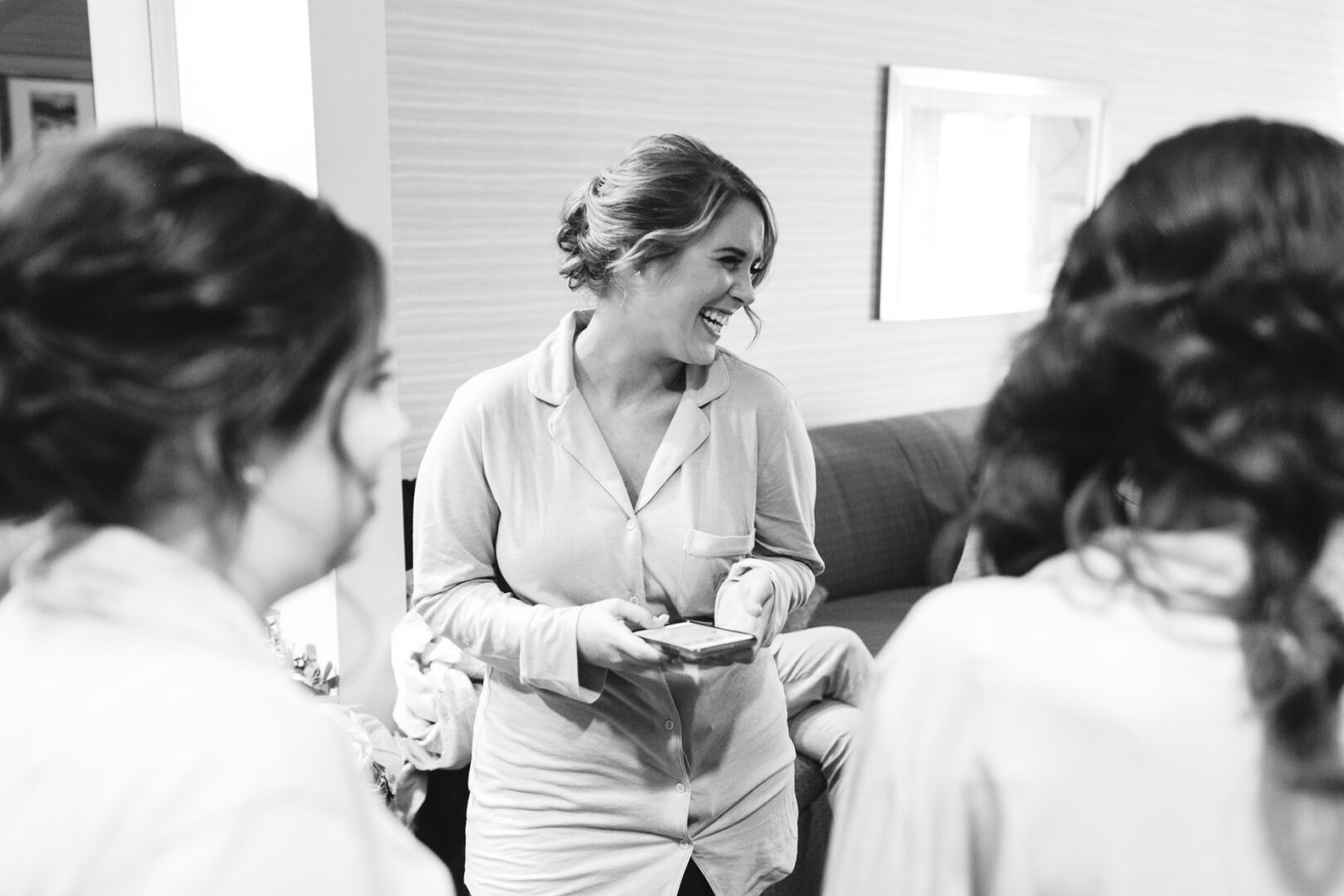 A woman in pajamas smiles while holding a phone, surrounded by friends in a casual indoor setting. The black and white image captures a moment as joyful as celebrating at the Palace at Somerset Park wedding.