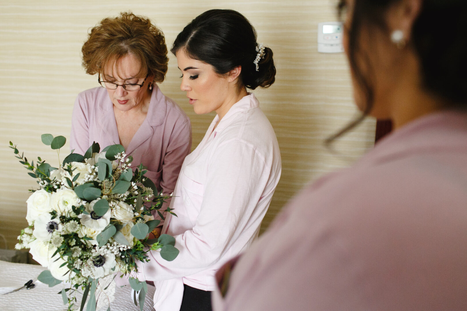 At a Palace at Somerset Park wedding, two women in pink outfits thoughtfully arrange a bouquet of white and green flowers, while another person is partially visible in the foreground.