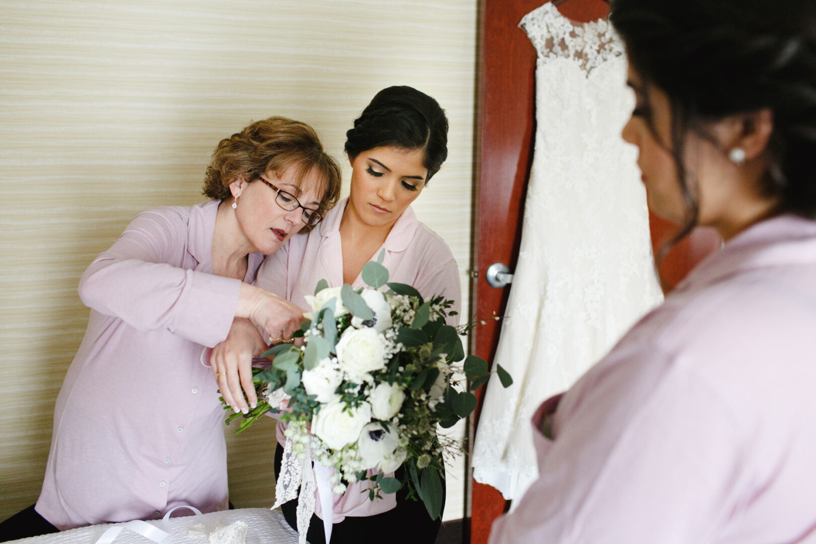 Two women in matching shirts delicately arrange a bouquet of white flowers, adding touches of greenery. In the background, a wedding dress graces the door, hinting at an elegant Palace at Somerset Park Wedding.