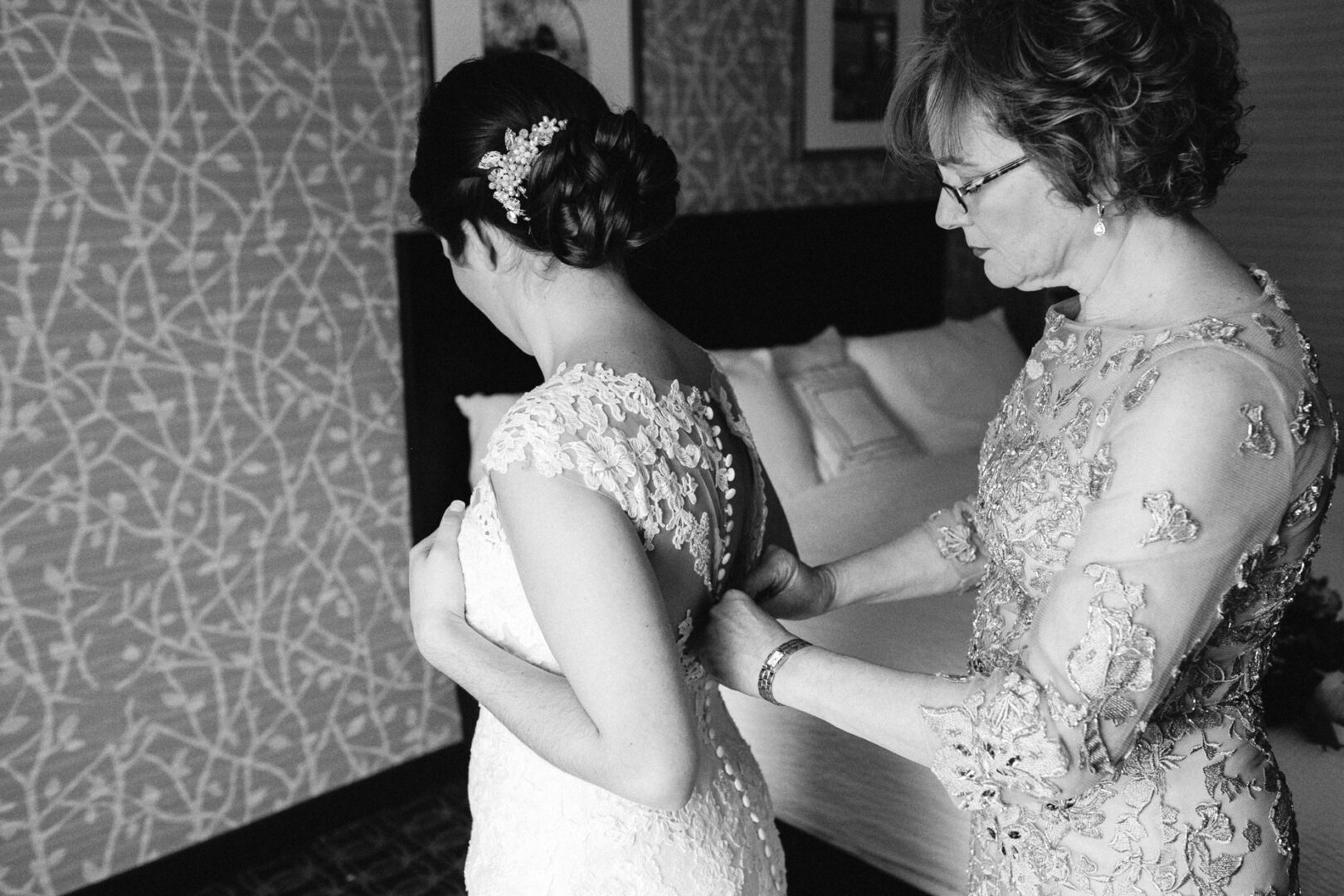 In a hotel room, a woman assists another in buttoning the back of her lace wedding dress, preparing for her Palace at Somerset Park wedding.