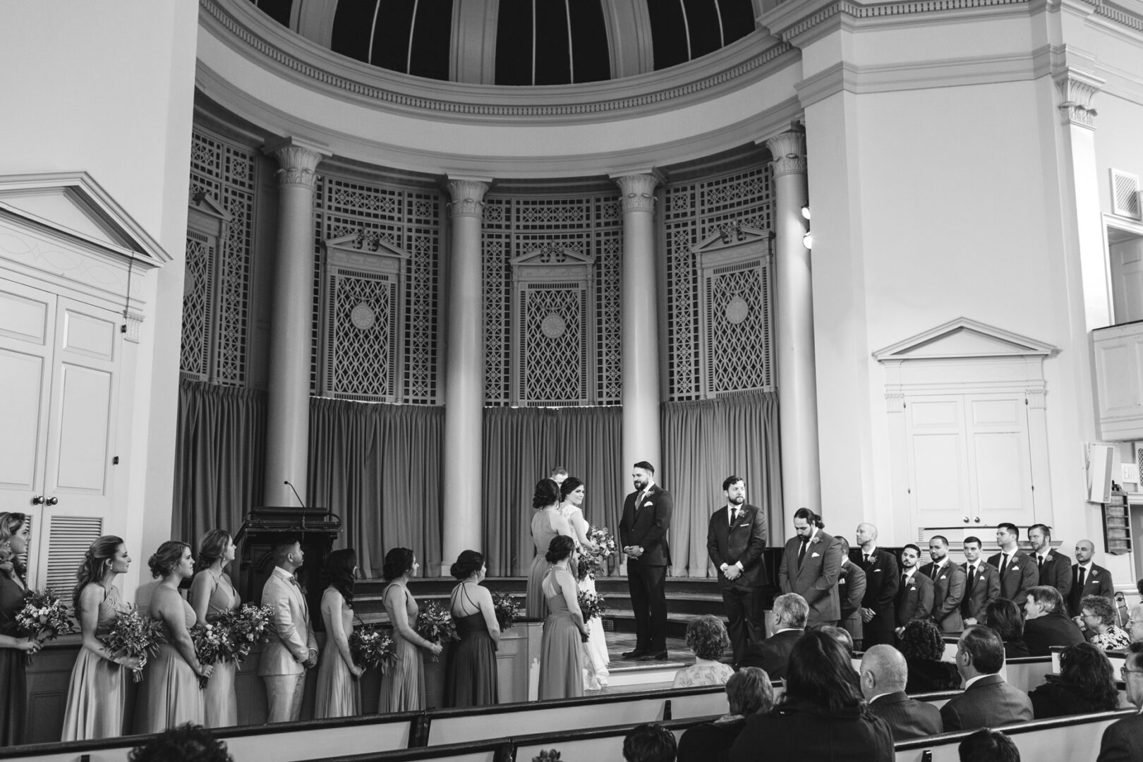 A black and white photo captures a Palace at Somerset Park wedding ceremony in a church. The bride and groom stand at the altar with bridesmaids and groomsmen on either side, as guests are seated, watching the poignant moment unfold.