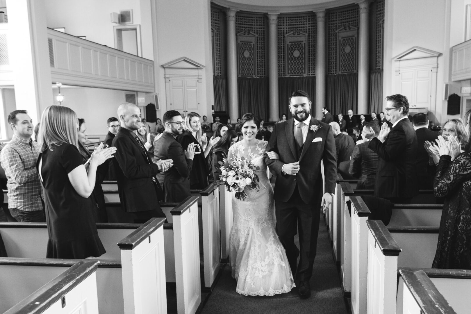 A bride and groom walk down the aisle of a church as guests clap, reminiscent of a grand Palace at Somerset Park wedding. The bride holds a bouquet, and the groom wears a suit.