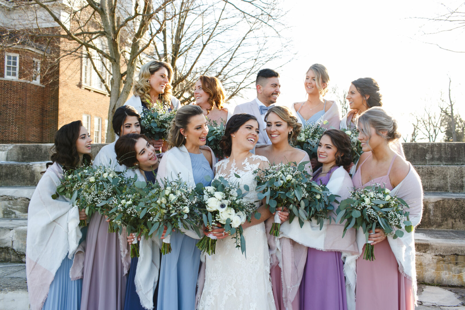 A bride and bridesmaids in pastel dresses and shawls stand outdoors, holding bouquets at their Palace at Somerset Park wedding. Amidst them, a man smiles warmly. In the background, elegant trees frame the scene alongside a charming brick building.