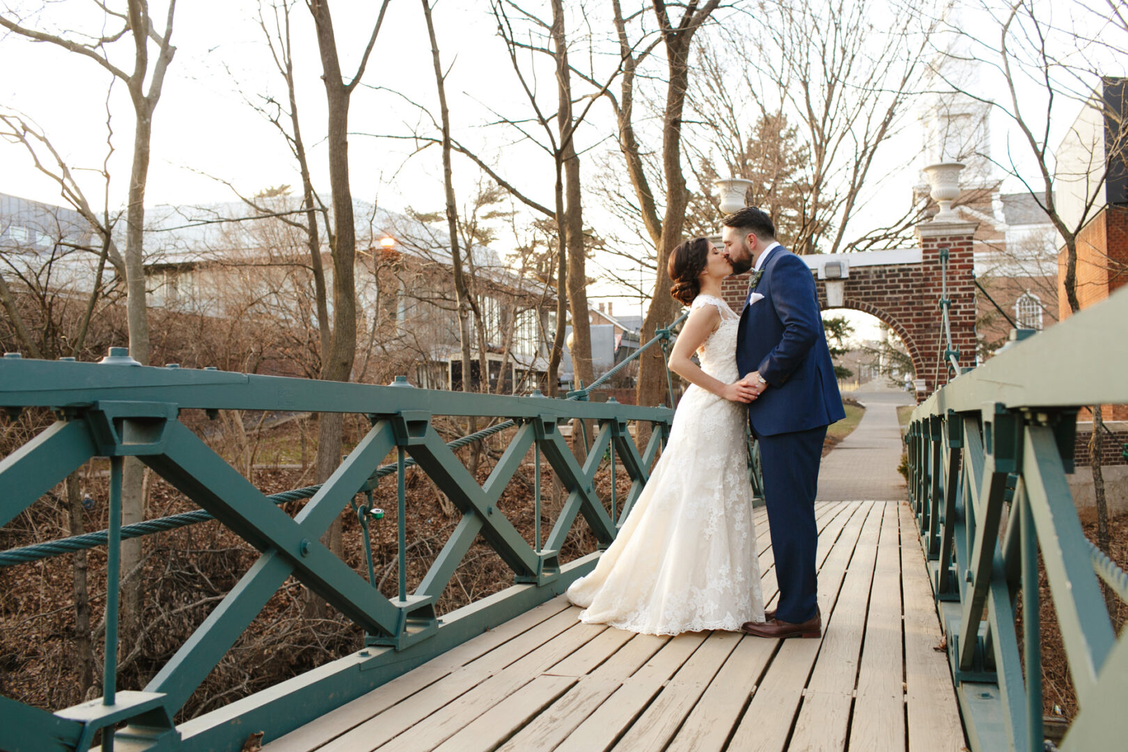 Bride and groom share a tender kiss on a wooden bridge, encircled by leafless trees with the elegant Palace at Somerset Park adding a touch of grandeur to the background.