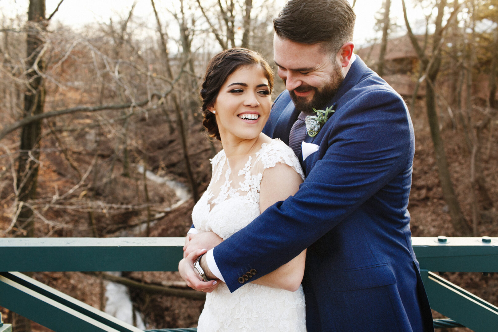 A couple in formal attire embraces on a bridge over a wooded area with a stream, capturing the enchanting essence of a Palace at Somerset Park wedding.