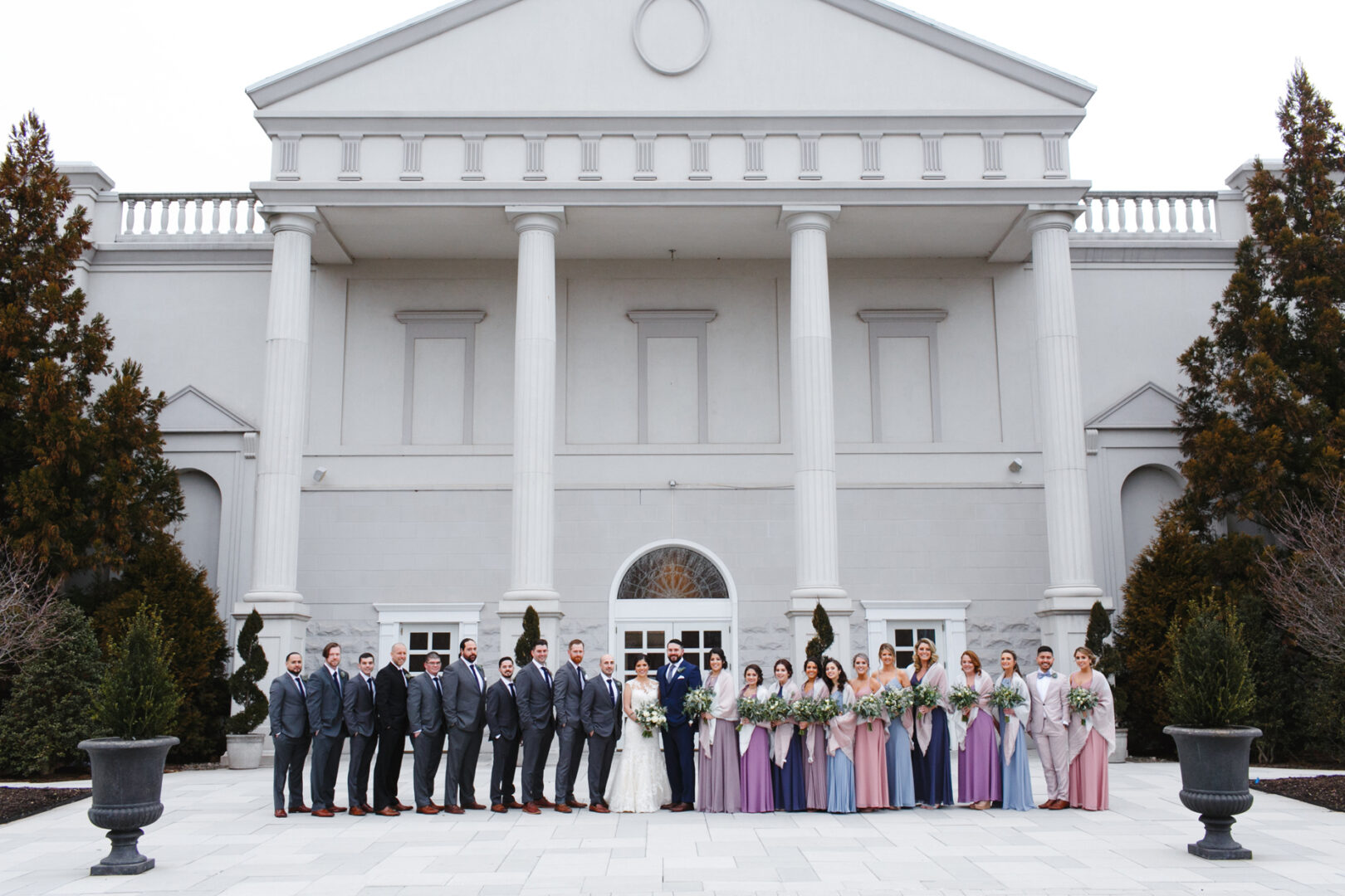 The wedding party poses elegantly in front of the grand Palace at Somerset Park, its tall white columns providing a majestic backdrop. The group, dressed in formal attire, features a radiant bride and groom celebrating their special day.