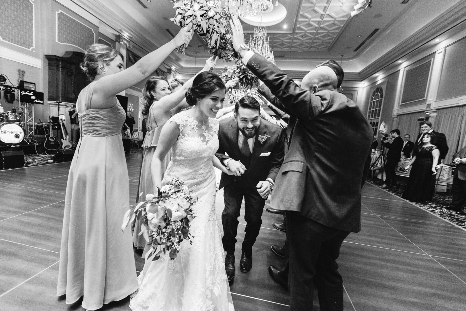 Bride and groom walk under a tunnel of raised arms made by guests on the dance floor at the Palace at Somerset Park. Guests cheer and smile in the elegant ballroom.
