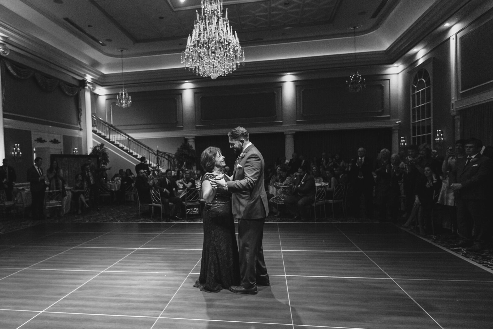 Amid the grandeur of the Palace at Somerset Park, a woman in a black dress and a man in a suit gracefully dance under the glistening chandelier. Guests watch in admiration from their tables, adding to the enchanting wedding atmosphere.