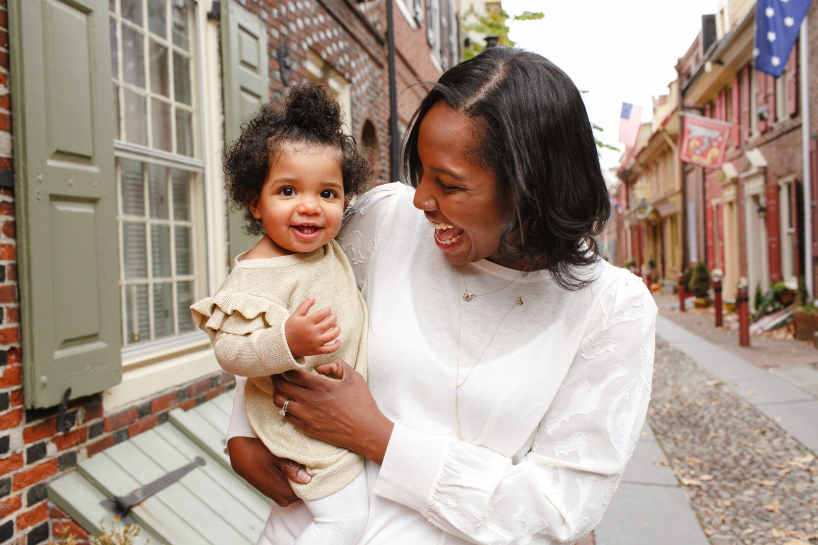 A woman smiles warmly at a baby she is holding on a brick-paved street lined with historic buildings, capturing the essence of Philadelphia family photos.