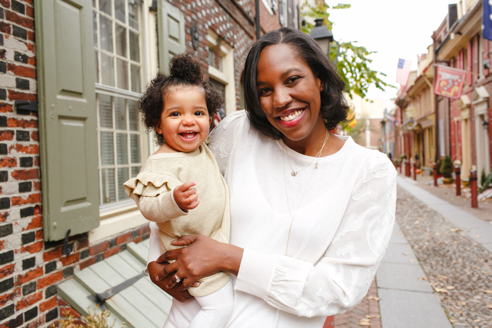 A woman holds a smiling child on a cobblestone street in Philadelphia, lined with historic brick buildings and flags, capturing a perfect family photo moment.