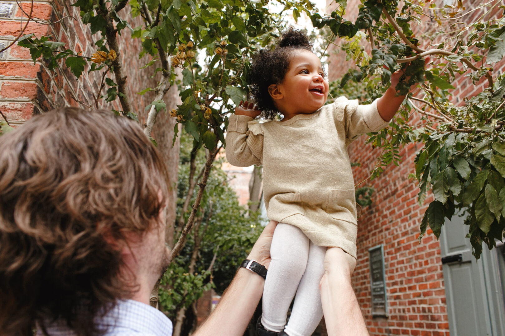 In the heart of a narrow brick alley, reminiscent of classic Philadelphia family photos, a person lifts a smiling child in a beige outfit towards the reaching tree branches above.