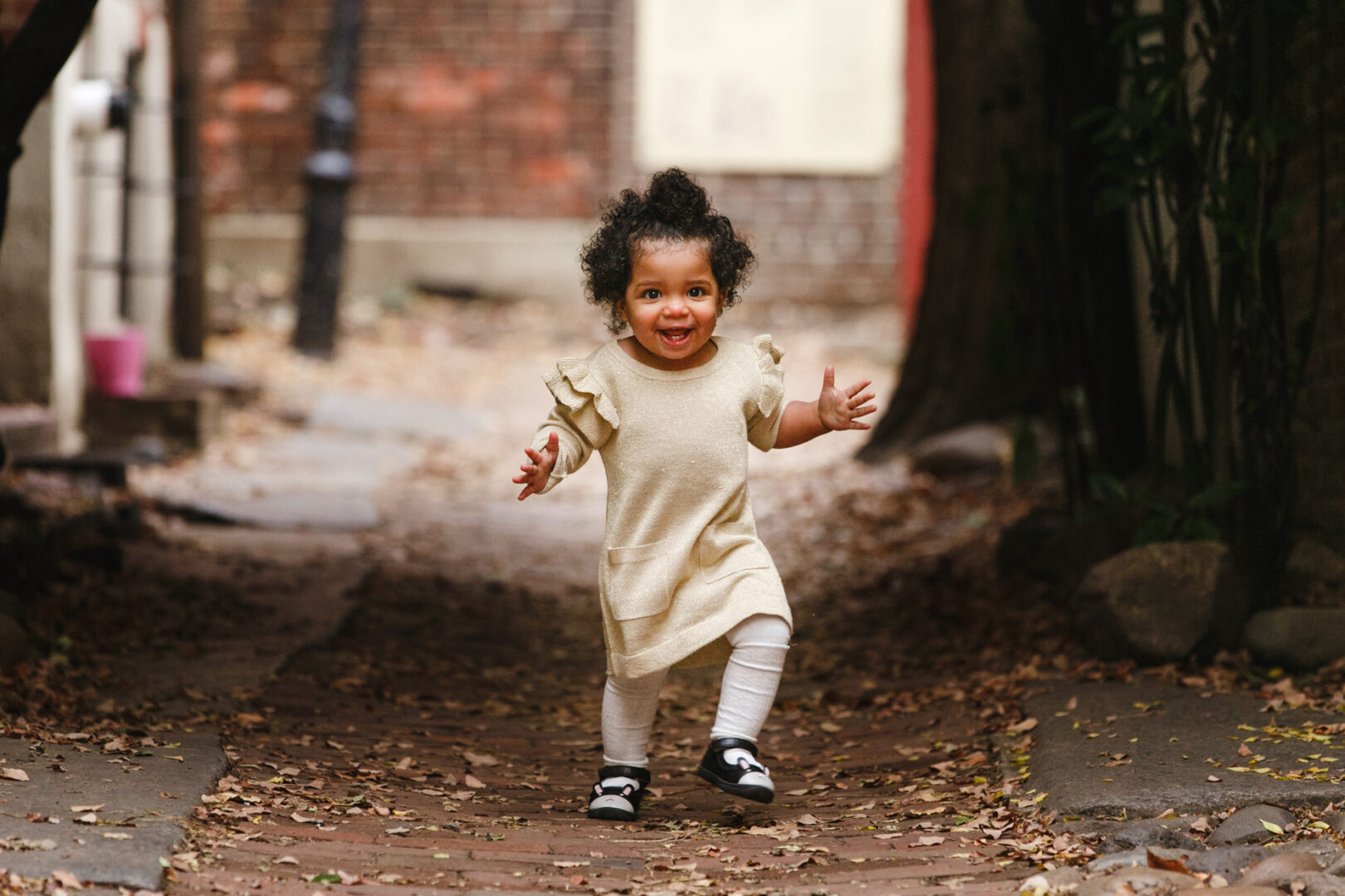 A young child with curly hair, wearing a beige dress and white leggings, joyfully runs down a leafy path, capturing the essence of Philadelphia family photos.