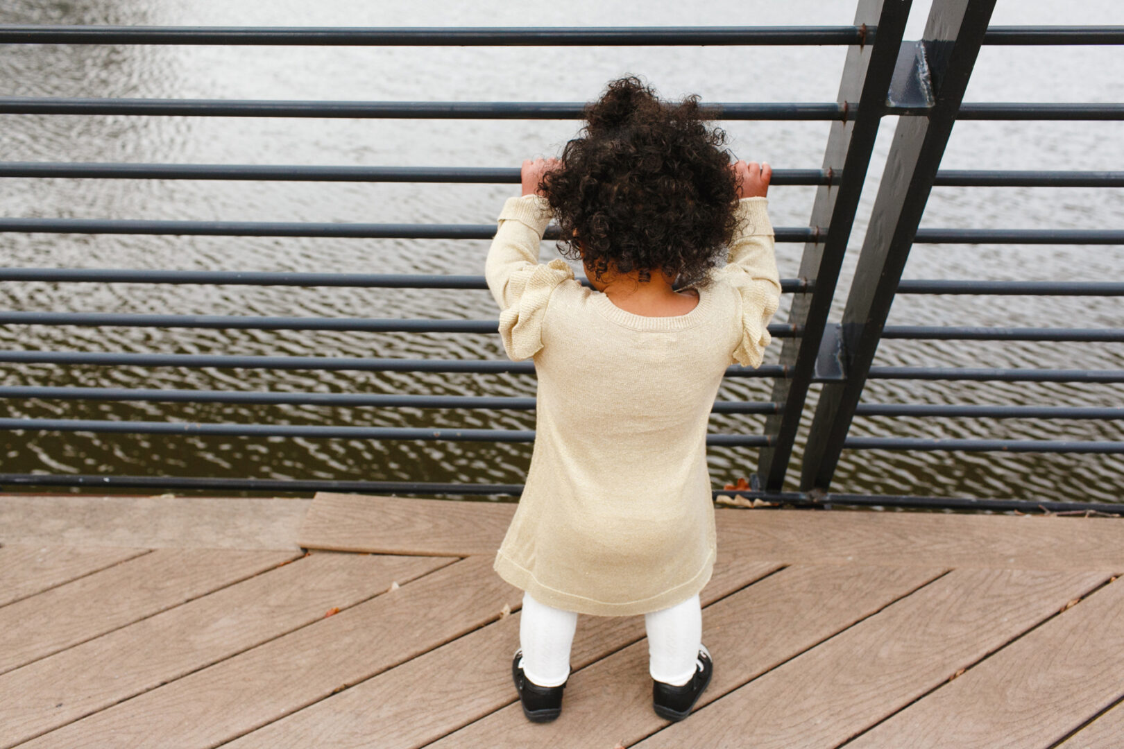 A child in a beige dress and white tights stands on a wooden deck, leaning against a metal railing while gazing thoughtfully at the water below. It's moments like these that capture the essence of Philadelphia family photos, where simple scenes tell timeless stories.