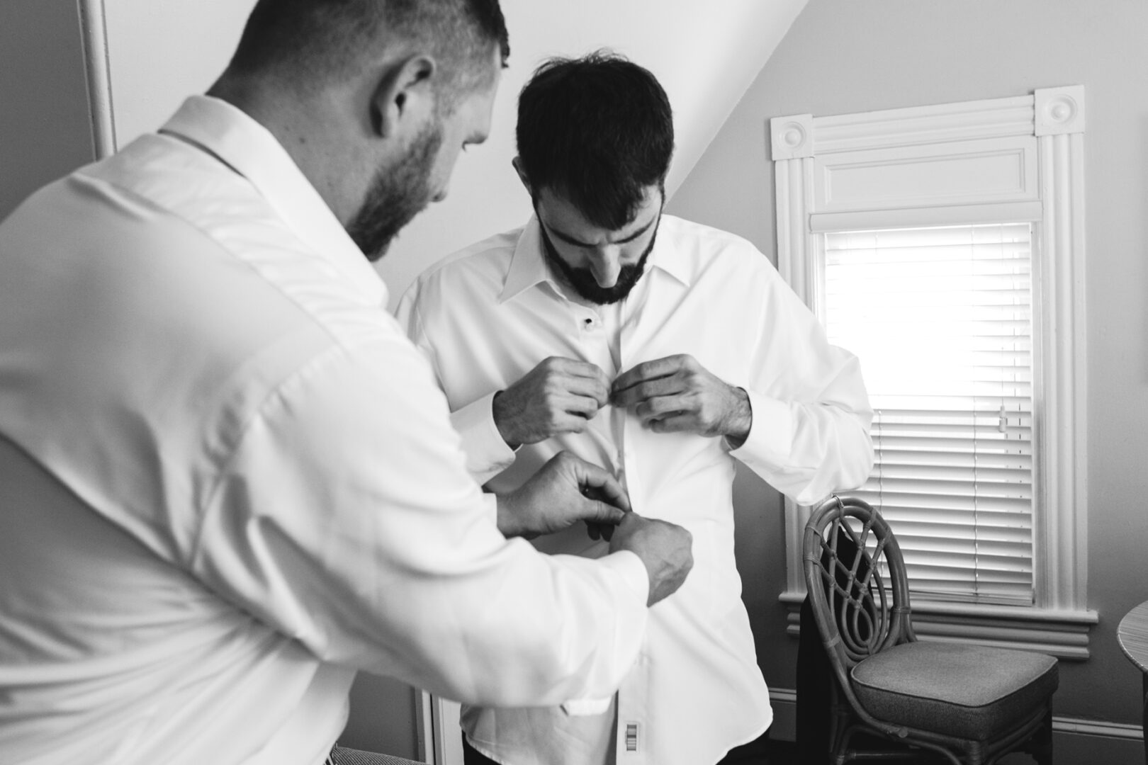 Two men in dress shirts adjust buttons on one man's shirt in a room with a window overlooking Willow Creek Winery. Black and white image.