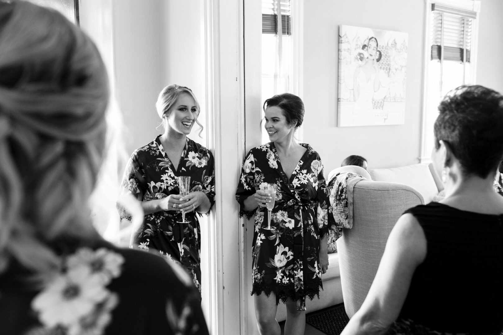 Two women in floral robes chat by a mirror, drinks in hand, as laughter fills the room. The ambiance reflects the charm of Willow Creek Winery, with elegant wall art and a cozy chair completing the scene. Two others stand nearby, their backs to the camera, absorbed in conversation.